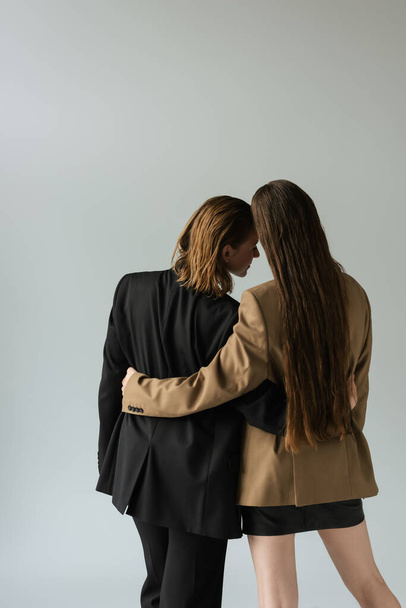 back view of woman with long hair hugging lesbian girlfriend in black blazer isolated on grey - Zdjęcie, obraz