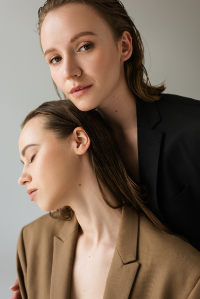 pretty young woman looking at camera near lesbian girlfriend with closed eyes isolated on grey - Fotoğraf, Görsel