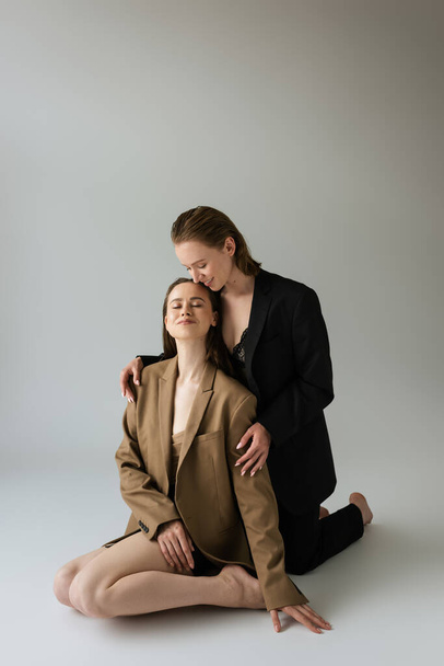 happy lesbian woman in black suit embracing girlfriend sitting with closed eyes on grey background - Foto, imagen