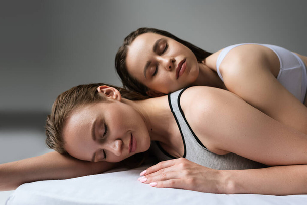 smiling woman with closed eyes relaxing on bed near brunette girlfriend on grey background - Foto, afbeelding
