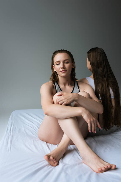 young lesbian woman sitting on bed with crossed legs near long haired girlfriend isolated on grey - Фото, изображение