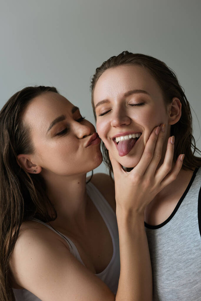 young woman pouting lips and touching face of lesbian girlfriend sticking out tongue isolated on grey - Foto, Bild