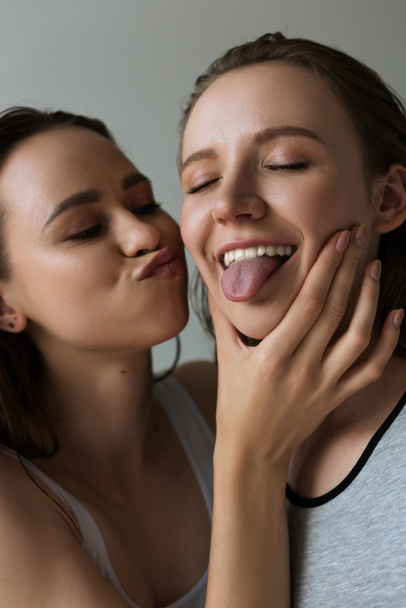 vrolijk vrouw met gesloten ogen steken uit tong in de buurt lesbisch vriendin pruilen lippen geïsoleerd op grijs - Foto, afbeelding