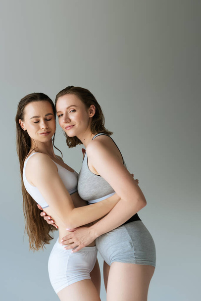 mladé lesbičky žena ve spodním prádle objímající brunetka přítelkyně a při pohledu na kameru izolované na šedé - Fotografie, Obrázek