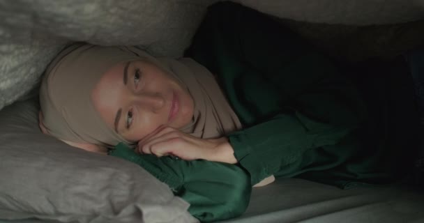 Jonge vrouw in een hijab ligt onder een deken. Dromerig glimlachend, knipperend. Een close-up. Hoge kwaliteit 4k beeldmateriaal - Video