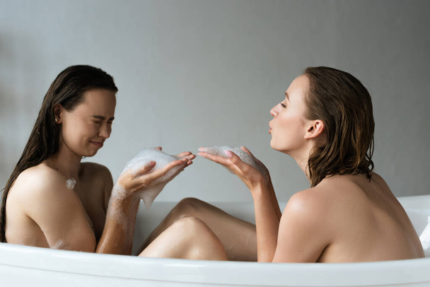 side view of nude lesbian couple having fun with soap foam in bathtub - Foto, afbeelding