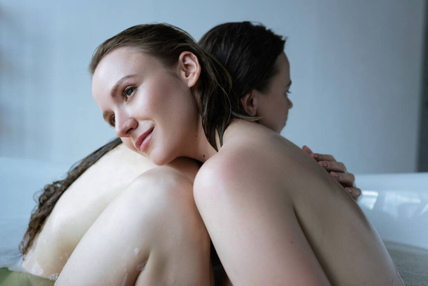 happy lesbian woman embracing with girlfriend in bathtub and looking away - Fotografie, Obrázek