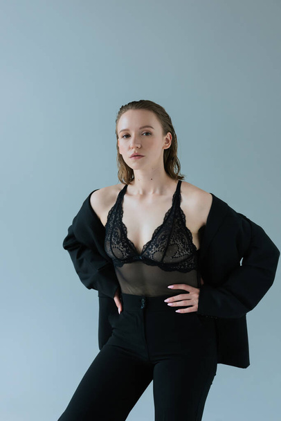 young woman in black trousers and black lingerie standing with hands on hips isolated on grey - Foto, afbeelding