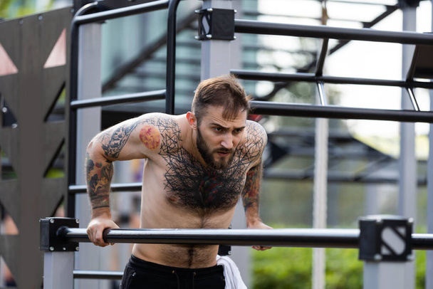 Homem bonito muscular está treinando parte superior do corpo em máquinas em campo de esportes de rua no centro da cidade - Foto, Imagem