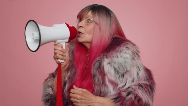 Smiling happy stylish old woman talking with megaphone, proclaiming news, loudly announcing sale advertisement, warning using loudspeaker to shout speech. Senior elderly grandmother on pink background - Footage, Video