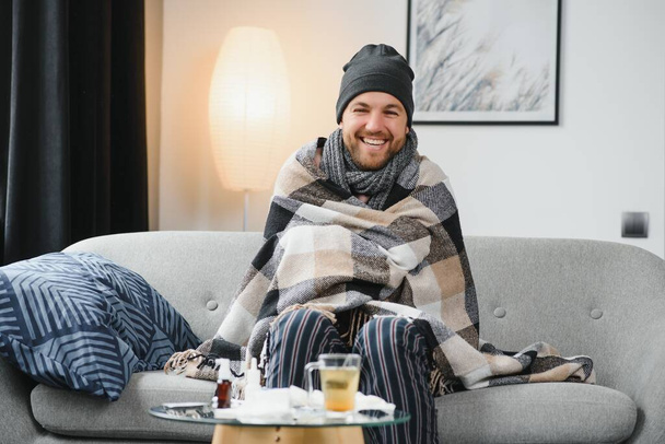 It's cold at home in wintertime. Man freezing in his house in winter because of broken thermostat. young guy wrapped in woolen plaid shivering while sitting on sofa in living room interior. - Фото, изображение