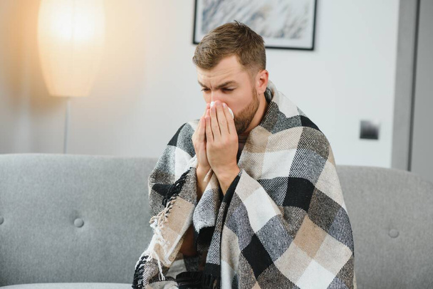 A sick man sits at home on a gray sofa with a blanket. Illness, protection, coronavirus, illness, flu. - 写真・画像