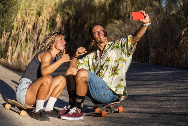 молода пара скейтбордистів відпочиває посміхаючись, приймаючи селфі зі своїм усміхненим телефоном, після сесії скейтбордингу на річковому каналі на заході сонця
. - Фото, зображення