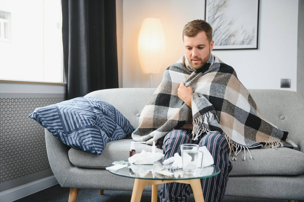 Sick bearded man who has bad cold or seasonal flu sitting on couch at home. Guy with fever wearing warm plaid shivering with worried face expression - Foto, Imagen