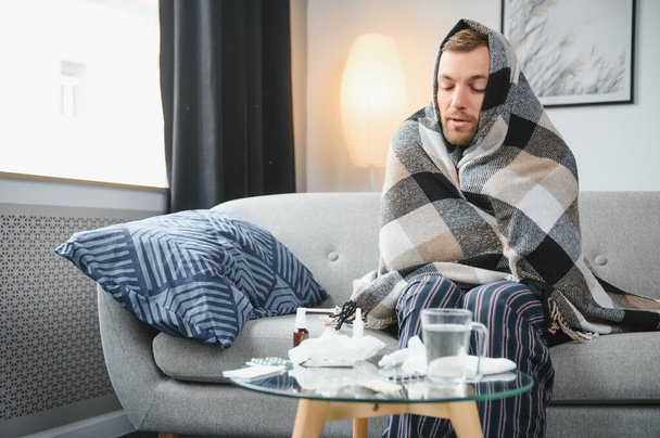Sick bearded man who has bad cold or seasonal flu sitting on couch at home. Guy with fever wearing warm plaid shivering with worried face expression - Foto, Imagen