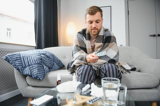 A sick man sits at home on a gray sofa with a blanket. Illness, protection, coronavirus, illness, flu. - 写真・画像