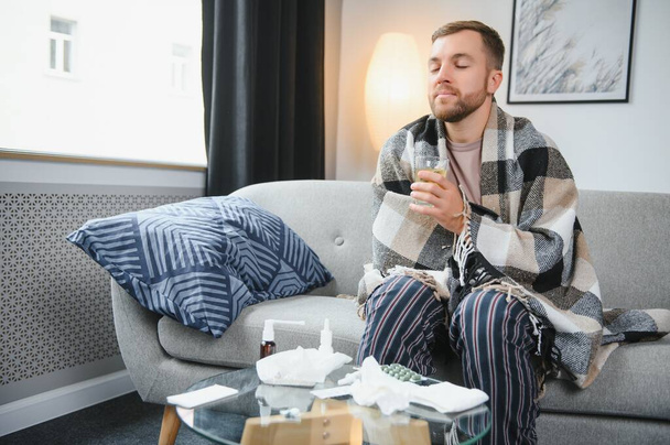 Sick bearded man who has bad cold or seasonal flu sitting on couch at home. Guy with fever wearing warm plaid shivering with worried face expression - Φωτογραφία, εικόνα