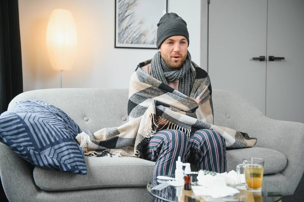 A sick man sits at home on a gray sofa with a blanket. Illness, protection, coronavirus, illness, flu. - Foto, afbeelding
