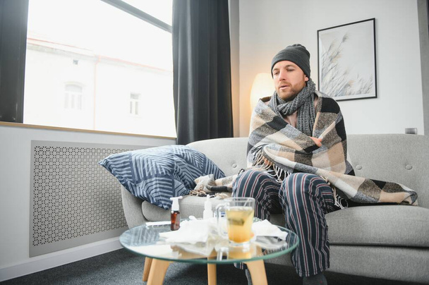 It's cold at home in wintertime. Man freezing in his house in winter because of broken thermostat. young guy wrapped in woolen plaid shivering while sitting on sofa in living room interior. - Fotografie, Obrázek