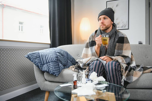 A sick man sits at home on a gray sofa with a blanket. Illness, protection, coronavirus, illness, flu. - Valokuva, kuva