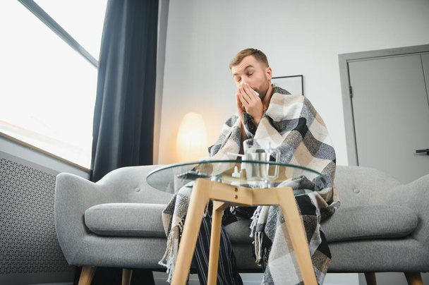 Sick sad man sits on couch at home suffers from runny nose flu disease coronavirus pandemic covid epidemic sneezes. Unwell guy feeling bad fever virus illness symptoms indoor - 写真・画像
