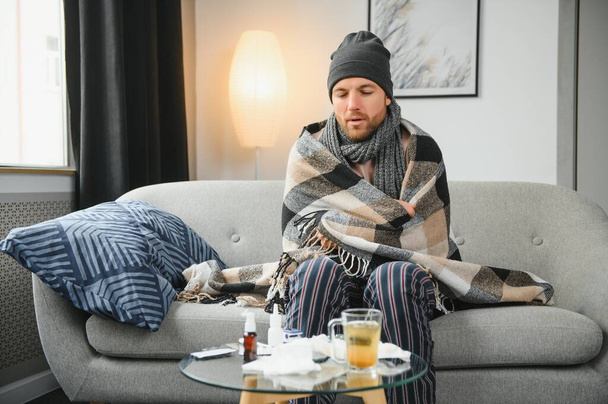 It's cold at home in wintertime. Man freezing in his house in winter because of broken thermostat. young guy wrapped in woolen plaid shivering while sitting on sofa in living room interior. - Photo, Image
