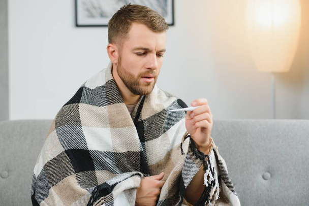 Sick adult man who has bad cold or seasonal flu virus is fighting illness at home. Unhappy middle aged man is sitting on sofa wrapped in warm scarf and plaid, holding paper tissue and sneezing - Foto, Bild