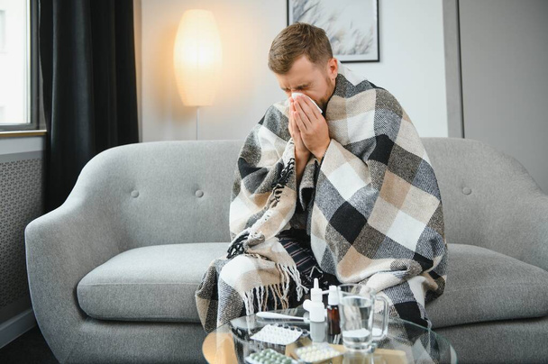 Person with cold blowing his nose. Sick man wearing scarf sitting on couch at home. Man with red running nose and cough. - Photo, Image