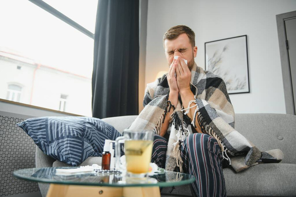 Nuori mies, joka kärsii flunssasta, flunssasta tai allergiasta, istuu kotona käärittynä peittoon ja pyyhkii nenänsä kudoksilla, kun hänellä on voimakas päänsärky kipu, terveydenhuollon käsite. - Valokuva, kuva