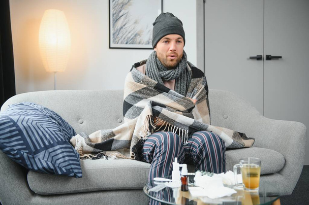 Sick bearded man who has bad cold or seasonal flu sitting on couch at home. Guy with fever wearing warm plaid shivering with worried face expression - Foto, immagini