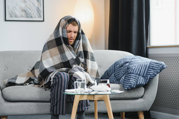 Sick adult man who has bad cold or seasonal flu virus is fighting illness at home. Unhappy middle aged man is sitting on sofa wrapped in warm scarf and plaid, holding paper tissue and sneezing - Foto, Imagen