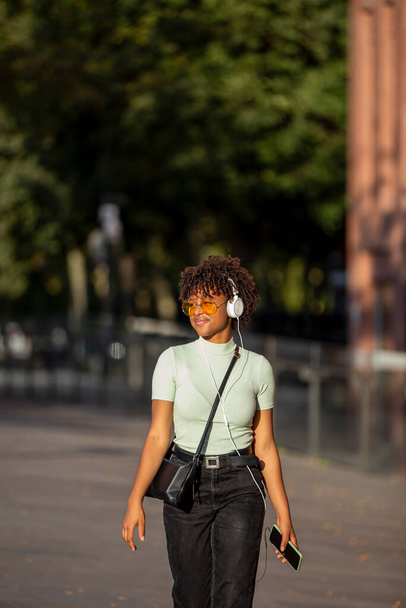 Stylowe ciemnoskóry kręcone młoda brunetka w żółtych okularach słonecznych cieszy się muzyką na słuchawki - Zdjęcie, obraz