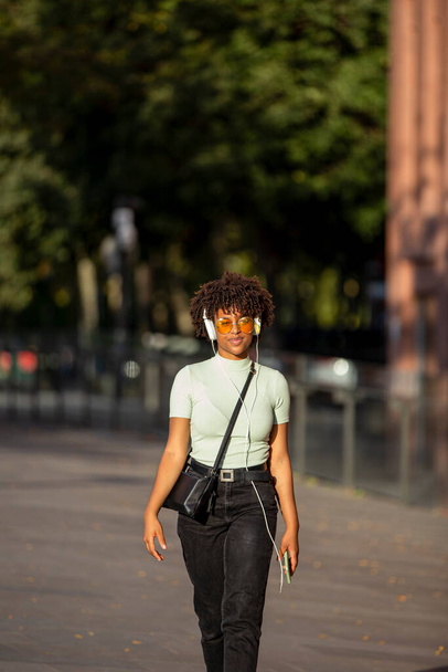 Stílusos sötét bőrű göndör, fiatal barna, sárga napszemüveges élvezi a zenét fülhallgatóban, miközben sétál az utcán - Fotó, kép
