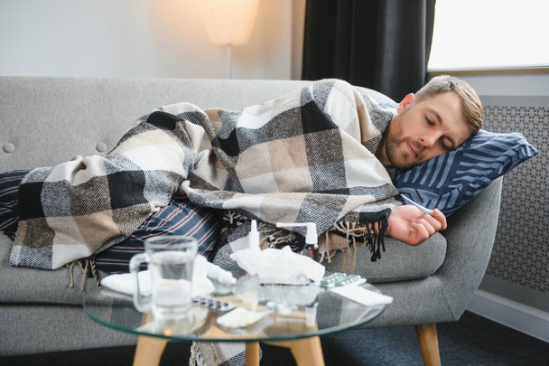 Sick bearded man who has bad cold or seasonal flu sitting on couch at home. Guy with fever wearing warm plaid shivering with worried face expression - 写真・画像