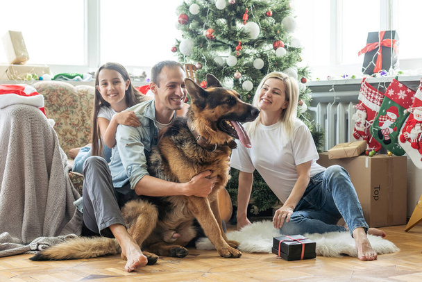 Ευτυχισμένη οικογένεια και χαριτωμένος σκύλος διασκεδάζουν στο χριστουγεννιάτικο δέντρο. ατμοσφαιρικές συναισθηματικές στιγμές. Καλά Χριστούγεννα και ευτυχισμένο το νέο έτος έννοια - Φωτογραφία, εικόνα