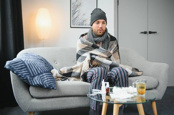 It's cold at home in wintertime. Man freezing in his house in winter because of broken thermostat. young guy wrapped in woolen plaid shivering while sitting on sofa in living room interior. - Fotografie, Obrázek