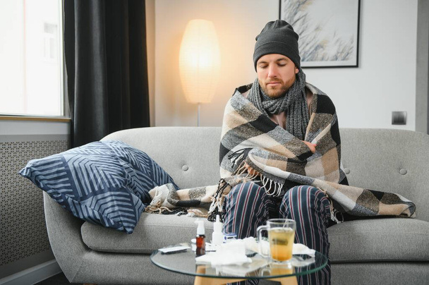 Sick bearded man who has bad cold or seasonal flu sitting on couch at home. Guy with fever wearing warm plaid shivering with worried face expression - 写真・画像