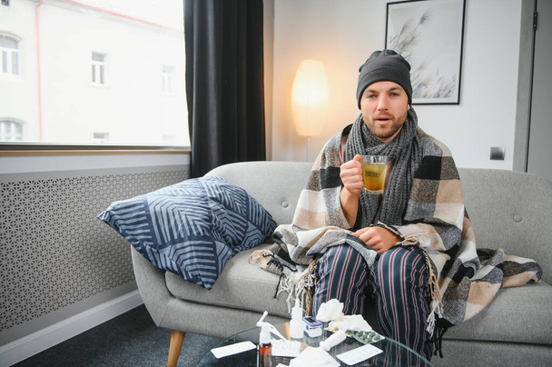 Young man suffering from a common cold and flu or allergy sit at home wrapped in blanket and wipe his nose with tissues while he have a strong headache pain, healthcare concept - Foto, Bild