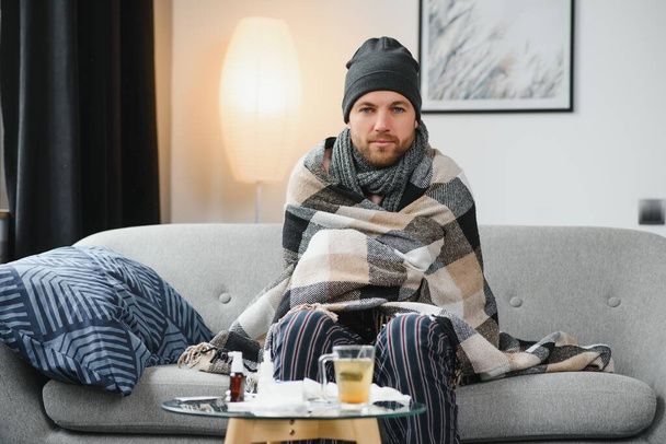 A sick man sits at home on a gray sofa with a blanket. Illness, protection, coronavirus, illness, flu. - Photo, image