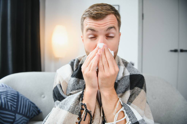 Person with cold blowing his nose. Sick man wearing scarf sitting on couch at home. Man with red running nose and cough. - Foto, Imagen