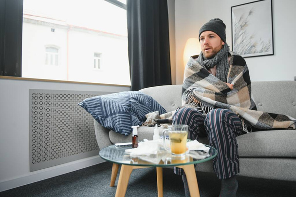 Sick bearded man who has bad cold or seasonal flu sitting on couch at home. Guy with fever wearing warm plaid shivering with worried face expression - Foto, Bild