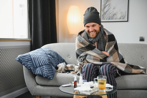 Sick bearded man who has bad cold or seasonal flu sitting on couch at home. Guy with fever wearing warm plaid shivering with worried face expression - Фото, изображение