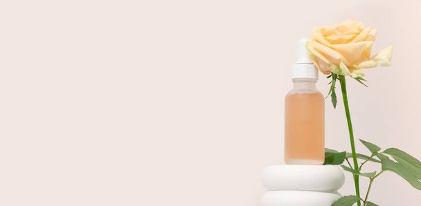 Kosmetik-Mockup-Serum mit Pipette in transparentem Milchglas und orangefarbener Flüssigkeit. Gesichtspeeling mit Milchsäure auf weißem Sockel, beiger Hintergrund mit Rosen - Foto, Bild