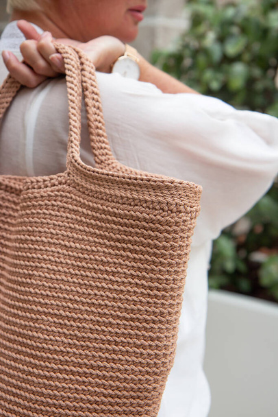 Woman holds a handmade beige knitted bag in her hand outdoors. Sustainable shopping. Wasteless lifestyle. Female with a jute bag with her own hands on a walk. Close up - Zdjęcie, obraz
