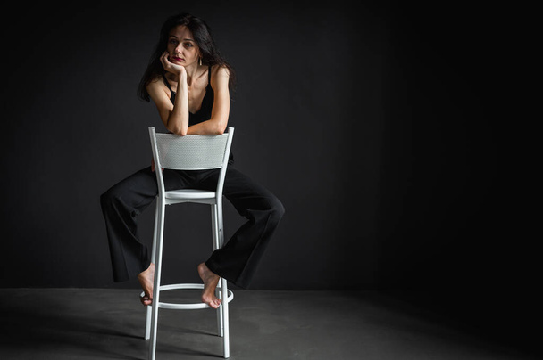 Fotografie celého těla roztomilé a svůdné brunetky žena sedí v kalhotách na židli izolované na tmavém pozadí  - Fotografie, Obrázek