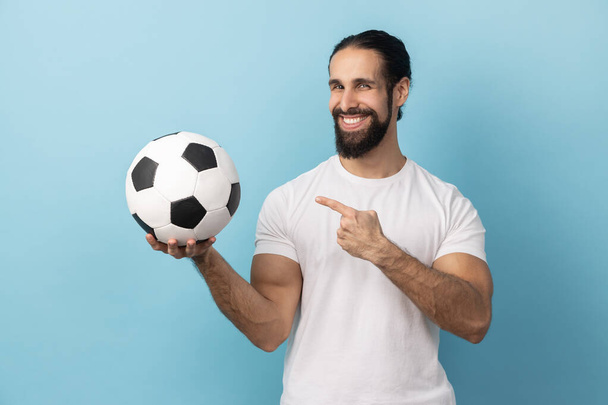Portrait of delighted man with beard wearing white T-shirt pointing at soccer ball in hand and looking at camera with toothy smile. Indoor studio shot isolated on blue background. - 写真・画像