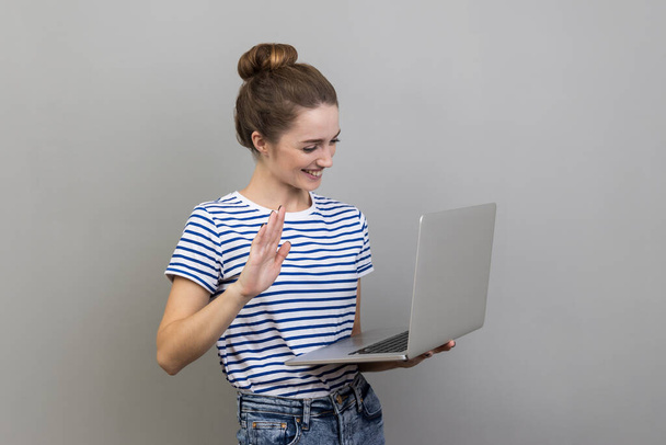 Portrait of friendly woman wearing striped T-shirt standing with laptop, looking at display and waving hand, saying hi, having video call. Indoor studio shot isolated on gray background. - Foto, Imagen