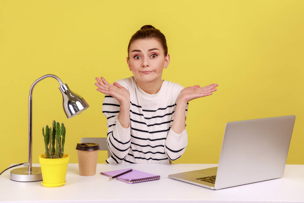 Confused uncertain woman office worker in striped shirt shrugging shoulders sitting on laptop at workplace, not sure about job completion. Indoor studio studio shot isolated on yellow background. - Φωτογραφία, εικόνα