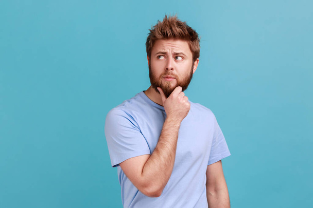 Portrait of unhappy pensive young bearded man keeping hand on chin, looking concentrated into distance, thinking about important things. Indoor studio shot isolated on blue background. - Фото, изображение