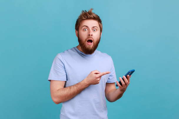 Portrait of astonished shocked bearded man pointing at cellphone and looking at camera with amazement, surprised about mobile data tariffs. Indoor studio shot isolated on blue background. - Photo, Image